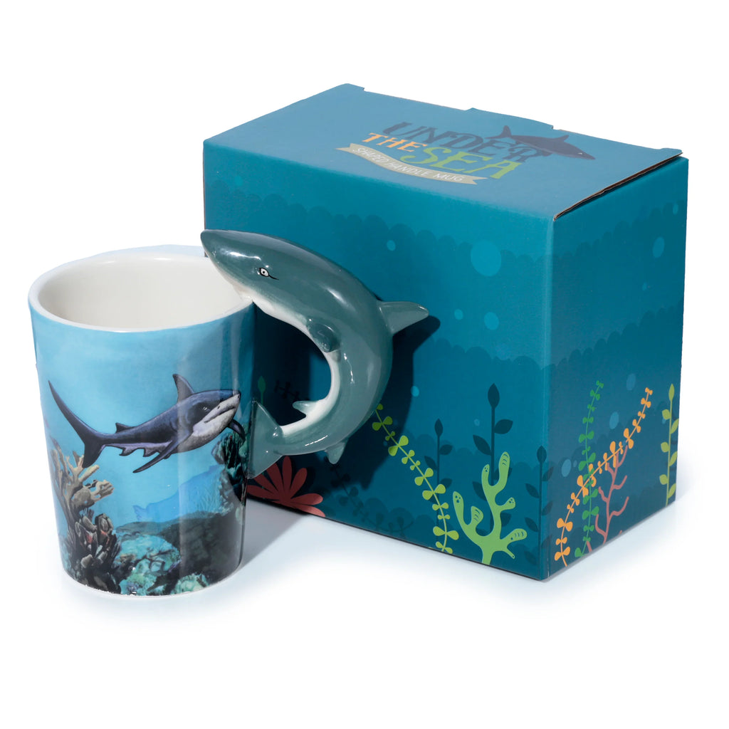 Angled shot displaying a ceramic mug with a detailed grey shark-shaped handle. The mug features an underwater scene with a large shark swimming amongst coral. Beside it is a teal blue gift box with 'Under the Sea' text and green and orange marine plant designs.
