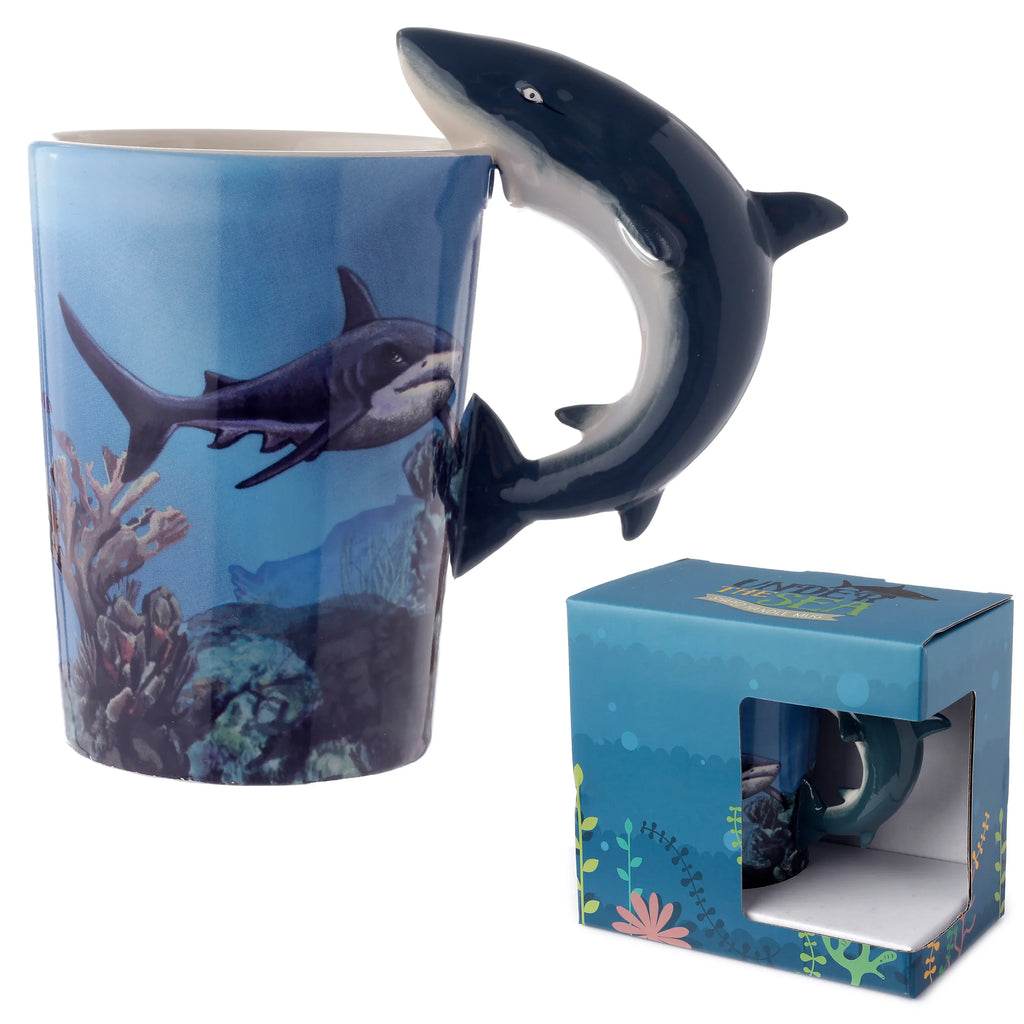 Angled view of a ceramic mug with a blue underwater design featuring a swimming shark and coral, complemented by a prominent dark blue shark-shaped handle. A blue marine-themed gift box is visible behind it.