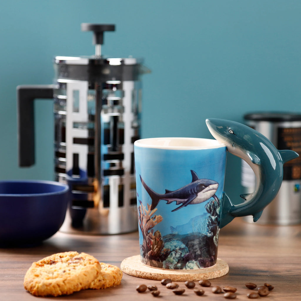 In-context lifestyle shot of a ceramic mug with a sculpted shark handle, displaying an underwater illustration of a shark and coral reef. The mug rests on a cork coaster, surrounded by coffee beans and cookies on a wooden table.