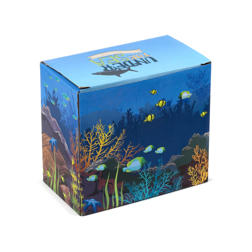 Angled view of a blue and dark blue ceramic mug packaging box. The box features an 'under the sea' design with colourful fish, coral, and seaweed, and the top displays a shark silhouette and 'UNDER THE SEA SHAPED HANDLE MUG' text.