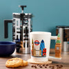 In-context lifestyle shot of a white ceramic Big Ben London Icons mug, showcasing its sculpted Big Ben handle and colourful illustrations of iconic London landmarks including Tower Bridge, The Gherkin, and a red double-decker bus, set on a wooden surface with a cork coaster, coffee beans, and biscuits.