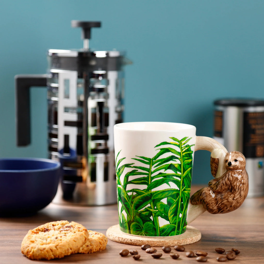 Angled lifestyle shot of the Jungle Explorer Sloth ceramic mug, featuring green jungle foliage and a brown shaped sloth handle, on a wooden table with coffee beans and biscuits.