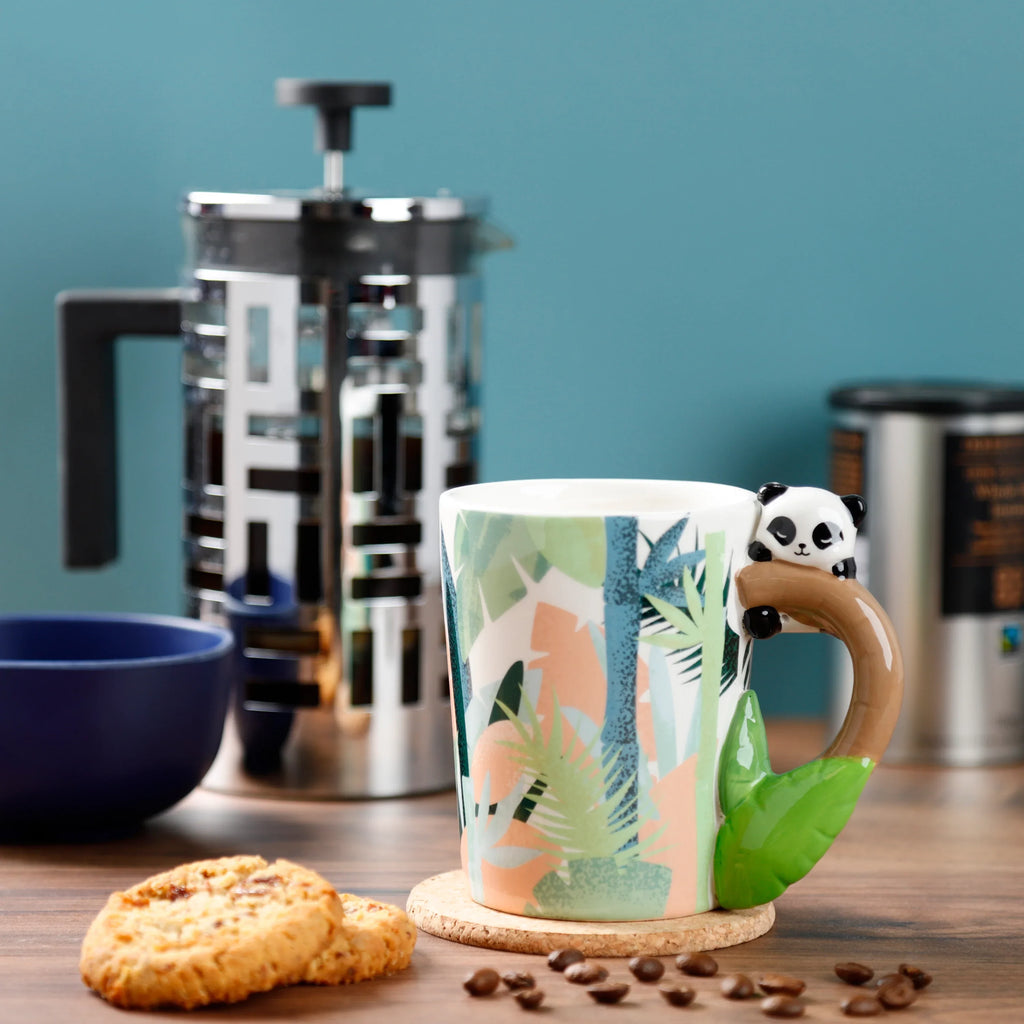 In-context lifestyle shot of a Pandarama Panda ceramic mug, featuring a tropical leaf pattern and a brown branch-shaped handle with a sculpted black and white panda. The mug is on a cork coaster, with biscuits and coffee beans nearby, and blurred kitchen items in the background.