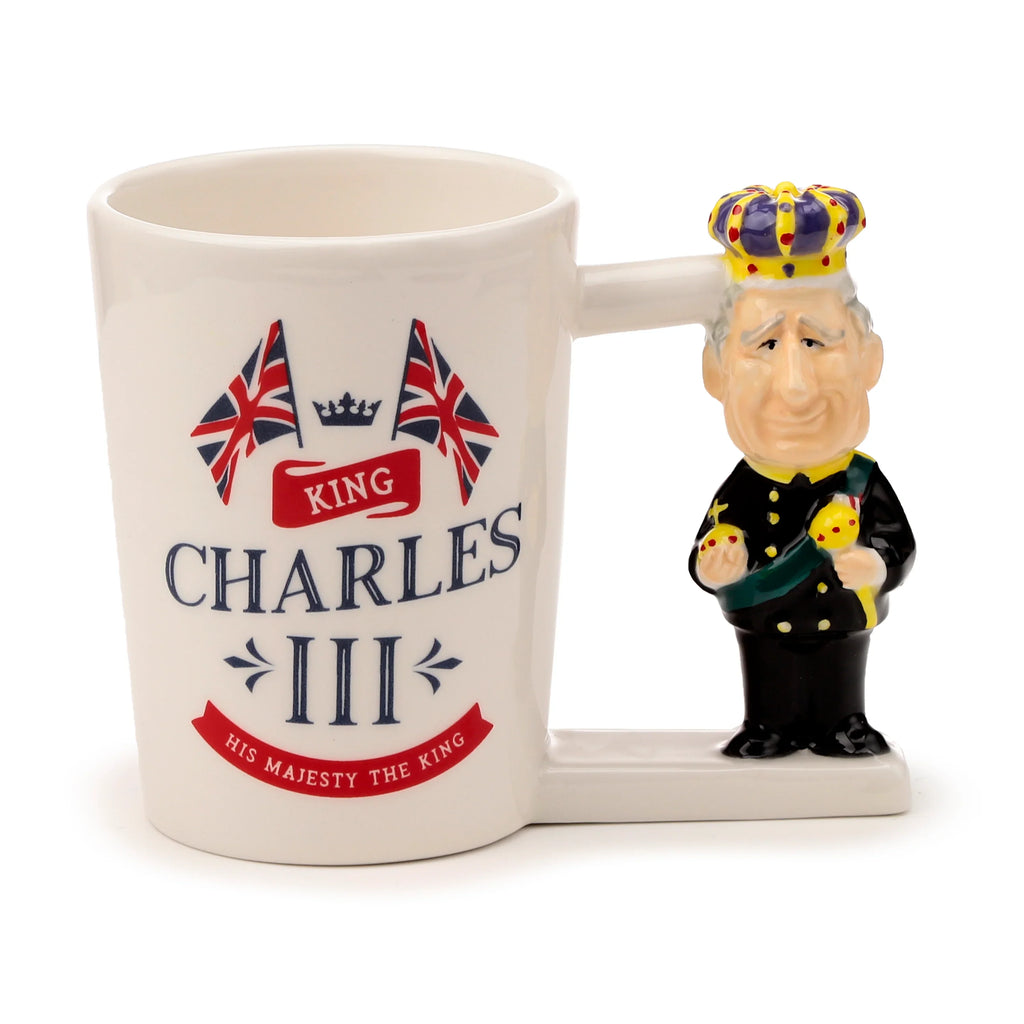 Angled view of a cream ceramic novelty mug, featuring a King Charles III shaped handle with a crown and uniform. The mug body is decorated with 'KING CHARLES III' text, Union Jack flags, and 'HIS MAJESTY THE KING' on a red banner.