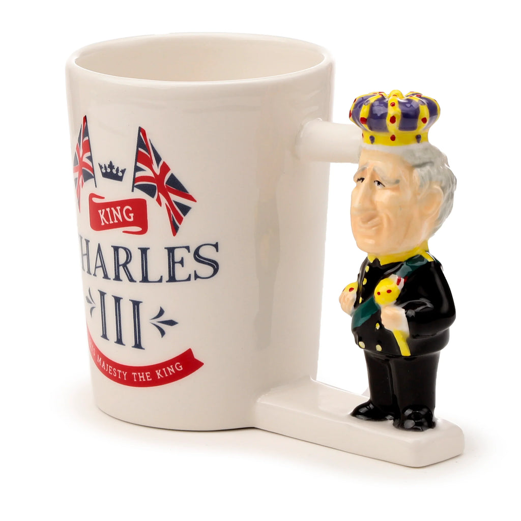 Angled view of a white ceramic King Charles III novelty mug. Its decorative handle is shaped like King Charles III, depicted in a black suit with a purple and yellow crown. The mug's body features 'King Charles III, His Majesty The King' text, flanked by Union Jack flags.
