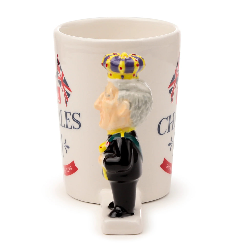 Side profile view of a cream ceramic novelty mug with a King Charles III shaped handle. The handle depicts the monarch in a black suit and colourful crown. Union Jack flag and 'CHARLES HIS KING' text are partially visible on the mug.