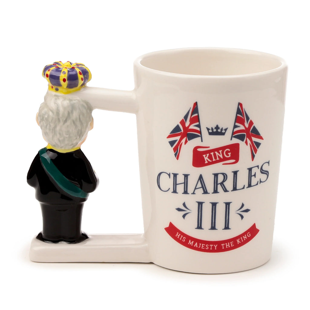 Angled view of a white ceramic novelty mug with a King Charles III shaped handle, visible from the rear. The handle features King Charles III wearing a black uniform, green sash, and a colourful crown. The mug body is decorated with two Union Jack flags and 'King Charles III, His Majesty The King' text.