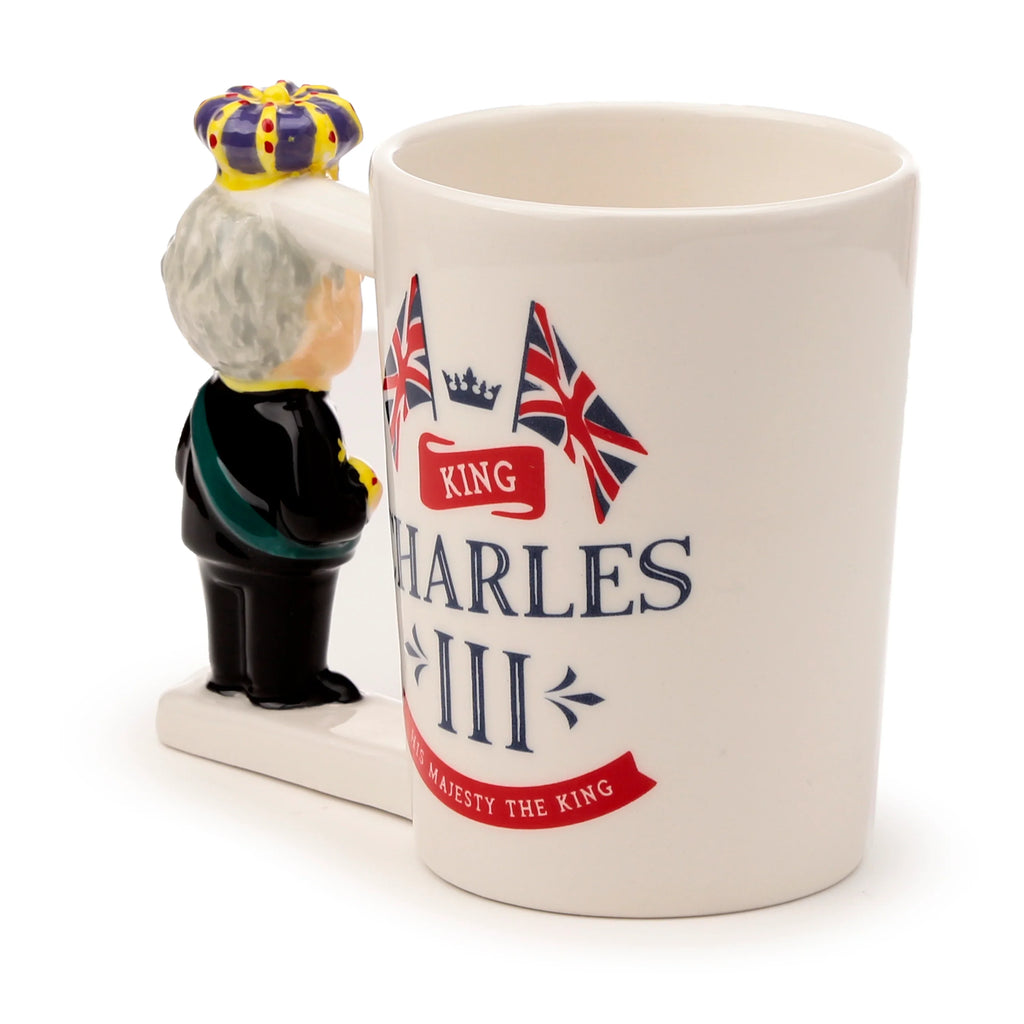 Angled view of a white ceramic novelty mug with a King Charles III shaped handle. The handle figure is seen from the back, wearing a crown and dark uniform. The mug is decorated with 'KING CHARLES III' text, 'HIS MAJESTY THE KING' on a red banner, a crown motif, and two Union Jack flags.