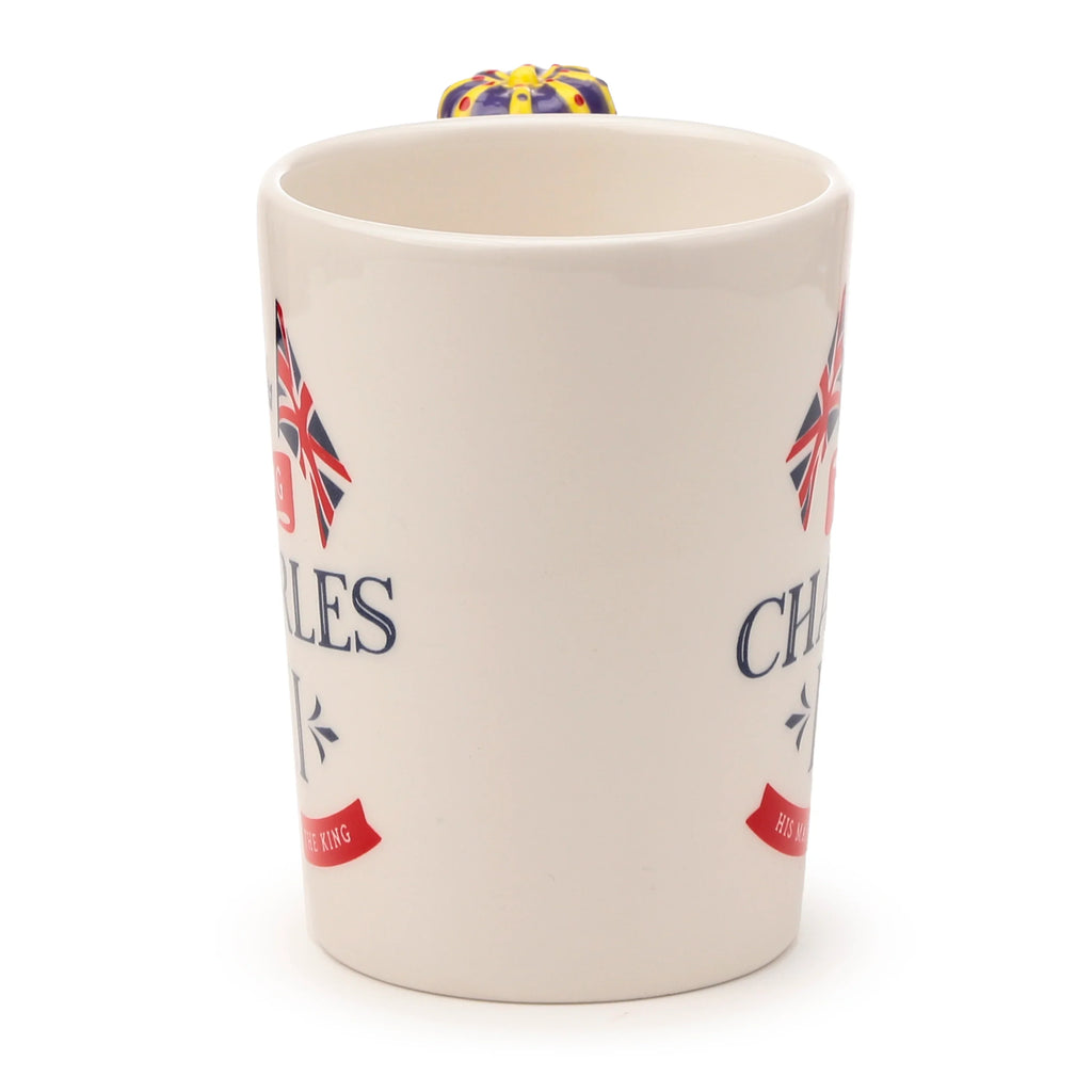 Cream ceramic novelty mug with a Union Jack flag design and 'CHARLES THE KING' text. The distinctive purple and yellow crown-shaped handle is prominently visible extending above the mug's rim, on a white background.