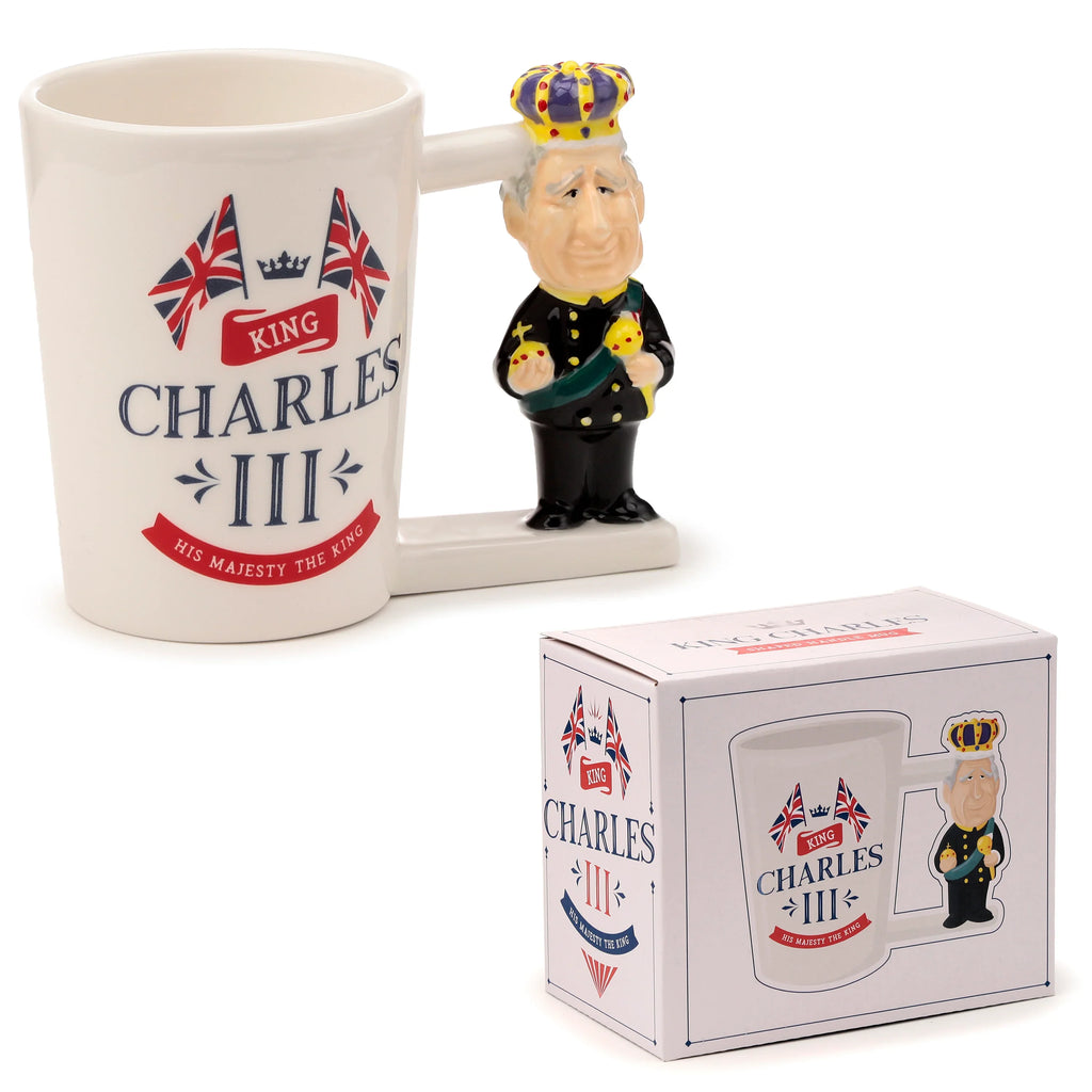 Angled view of a cream novelty ceramic mug with a King Charles III shaped handle, featuring Union Jack flags and 'King Charles III' text, presented alongside its illustrated packaging box.