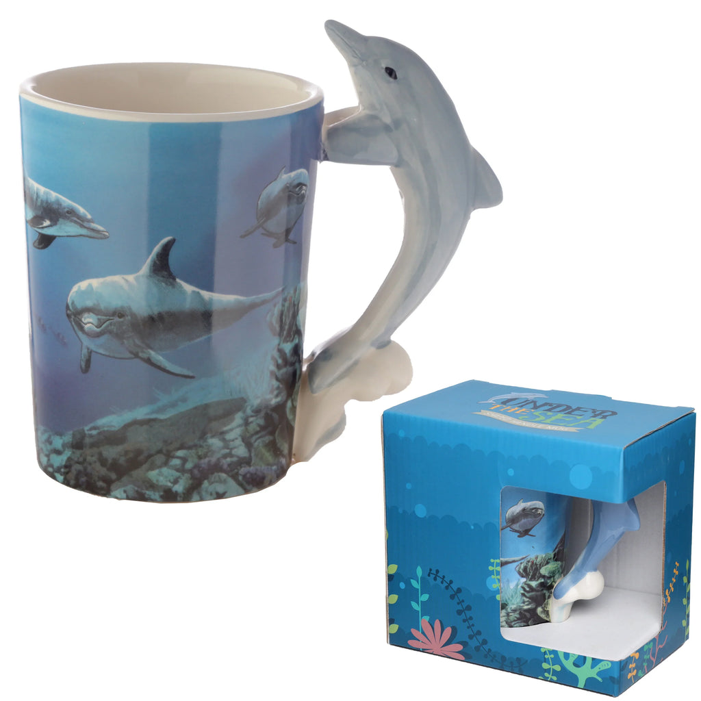 Angled view of a ceramic mug featuring an underwater scene with multiple dolphins on a blue background, complemented by a realistic, sculpted dolphin handle. The mug is presented alongside its blue display packaging.