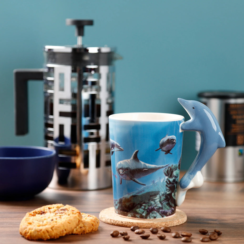 In-context lifestyle shot of a ceramic Dolphin Underwater Decal Mug with a blue shaped dolphin handle, angled to show its underwater scene print. The mug sits on a cork coaster on a wooden table, alongside two cookies and scattered coffee beans.