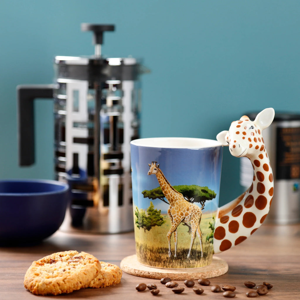 In-context lifestyle shot of a ceramic Giraffe Savannah Decal Mug, angled to prominently display its unique shaped giraffe handle and a decal of a tall giraffe in a savannah landscape. The mug rests on a cork coaster on a wooden surface, accompanied by coffee beans and biscuits, with a cafetière and blue bowl in the blurred background.