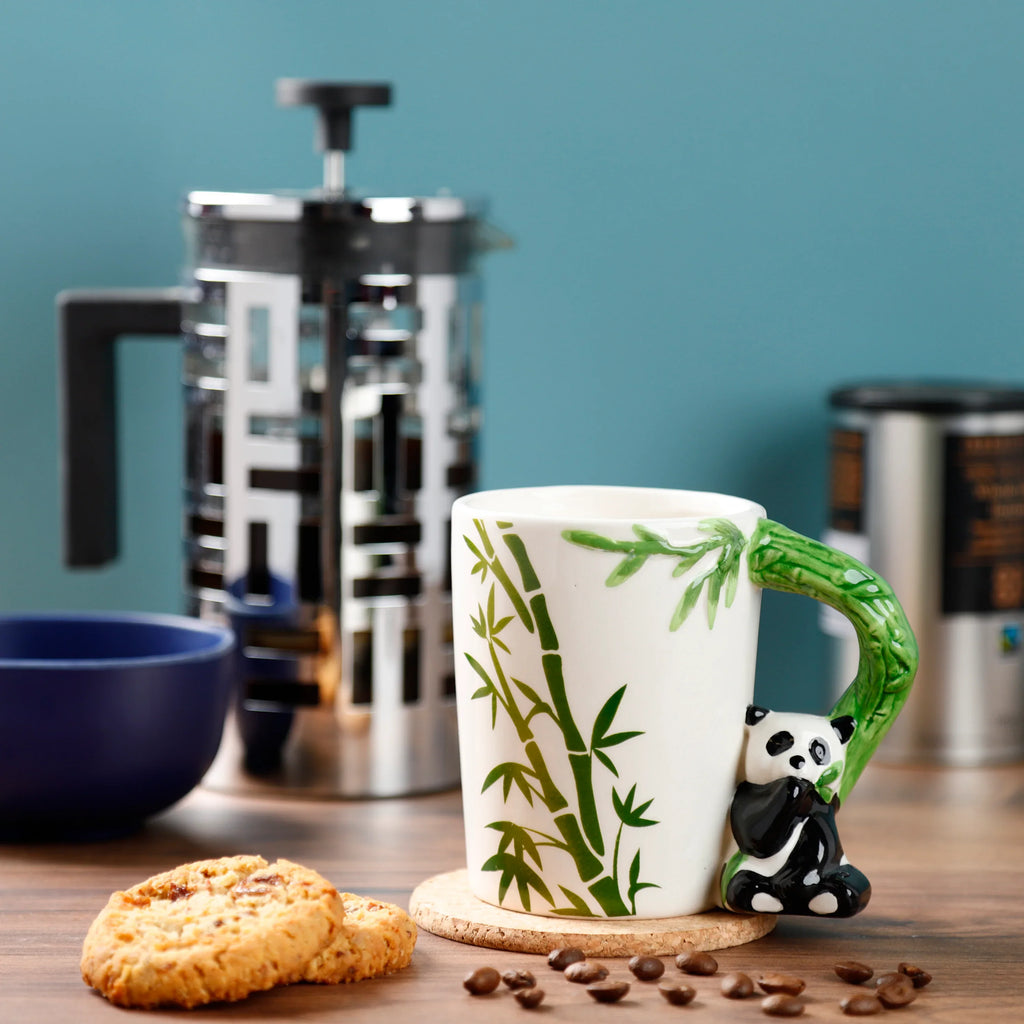 In-context lifestyle shot of a white ceramic mug with a green bamboo decal, an angled view revealing its shaped green bamboo handle and an attached black and white panda figure, set on a wooden surface with coffee beans and biscuits.