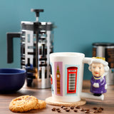 In-context lifestyle shot of a white ceramic Queen mug with a sculpted queen handle in purple and gold. The mug displays British-themed illustrations of a Queen's Guard, Big Ben, and a red telephone box. It rests on a cork coaster on a wooden table, surrounded by cookies and scattered coffee beans.