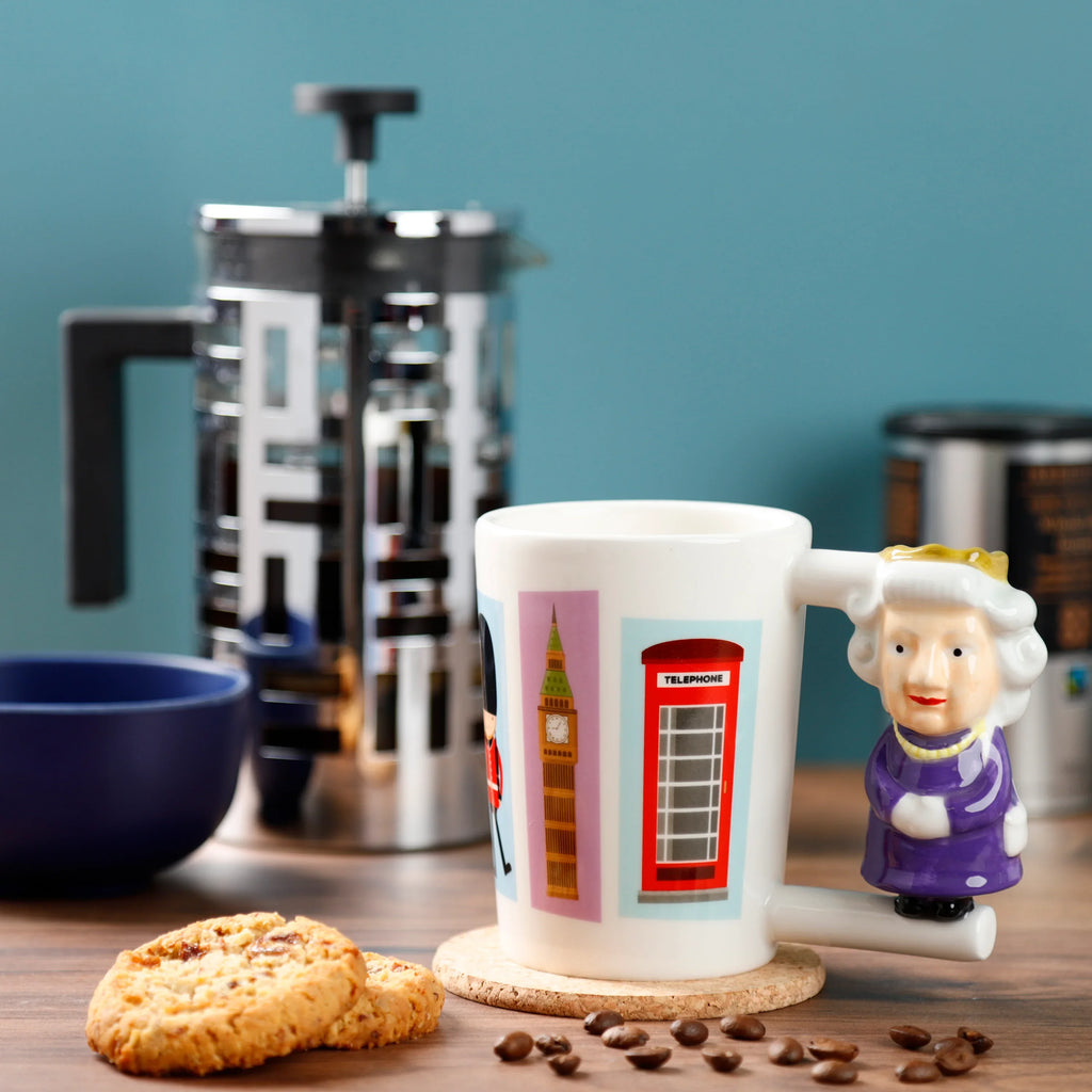 In-context lifestyle shot of a white ceramic Queen mug with a sculpted queen handle in purple and gold. The mug displays British-themed illustrations of a Queen's Guard, Big Ben, and a red telephone box. It rests on a cork coaster on a wooden table, surrounded by cookies and scattered coffee beans.