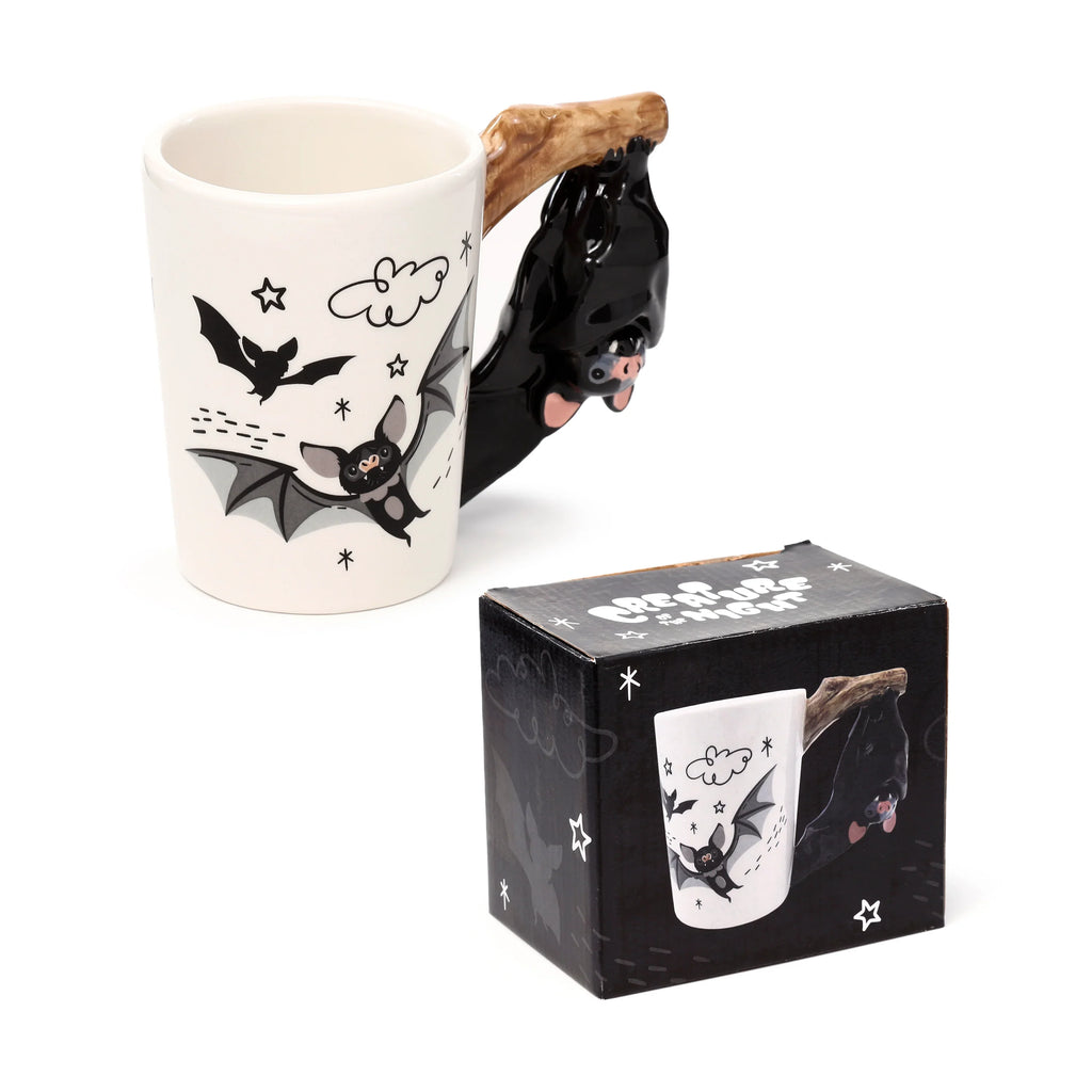 Angled product shot showing a cream ceramic mug with a shaped handle, depicting a black bat hanging from a brown branch. The mug's body is decorated with black and grey cartoon bats, clouds, and stars. A black coordinating gift box with similar bat graphics is positioned beside it.