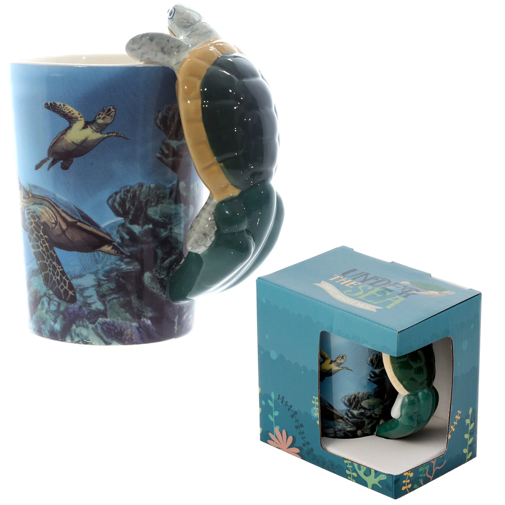 Angled view of a blue ceramic mug with a green and beige turtle-shaped handle and an underwater decal depicting turtles and coral, displayed next to its coordinating blue retail box with a clear window.