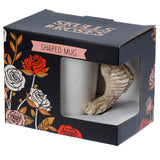 Angled view of a white ceramic mug with a beige skeleton-shaped handle, presented in its dark grey retail packaging. The box features red, white, and pink rose illustrations and 'SKULLS & ROSES' text.