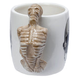 Close-up, angled view of a white ceramic mug, prominently featuring a detailed, light brown skeleton-shaped handle. A partial black skull graphic is visible on the mug's side.