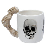 Angled view of a white ceramic mug with a bone-coloured, sculpted skeleton-shaped handle and a monochrome skull graphic on its side.