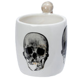 Angled view of a white ceramic mug with a black and white skull graphic, accented with subtle golden colour, and a light brown, skeleton-shaped handle.