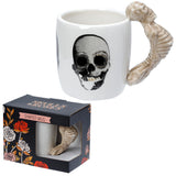 Angled view of a white ceramic skeleton mug with a prominent black skull graphic and a bone-coloured skeleton-shaped handle, shown next to its black packaging box featuring a rose design.