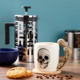 In-context shot of a cream ceramic mug with a black and white skull print and a sculpted skeleton handle, on a cork coaster with coffee beans and cookies on a wooden surface.