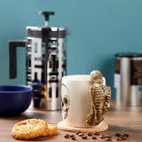 Cream ceramic Skeleton Shaped Handle Mug featuring a brown skeleton handle and a black skull design, shown in a lifestyle shot on a cork coaster on a wooden table with scattered coffee beans and two biscuits.