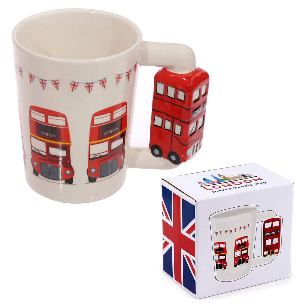 Angled view of a white ceramic Routemaster London bus mug with a red London bus-shaped handle, decorated with red bus illustrations and Union Jack bunting. A white retail box displaying a Union Jack flag and mug graphic sits alongside.