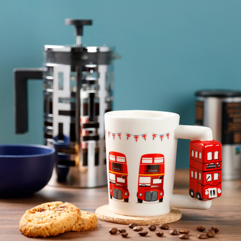 Angled lifestyle shot of a white ceramic mug with red double-decker London bus illustrations and a red double-decker bus shaped handle, displayed on a wooden surface with coffee beans and biscuits.