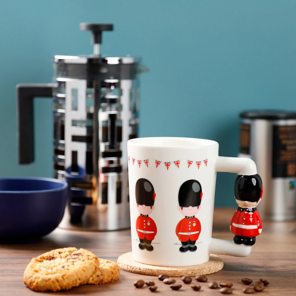 In-context lifestyle shot of a white ceramic Guardsman Figure shaped handle mug, decorated with red guardsmen and Union Jack bunting, sitting on a cork coaster with scattered coffee beans and biscuits.