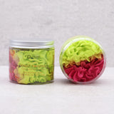 Front view of an upright transparent whipped soap jar with a silver-coloured lid, displaying layers of bright green, orange, and fuchsia whipped soap, alongside its 'Kiwi & Strawberry Scented' label. To its right, a second transparent jar is angled to showcase a close-up of the vibrant green, orange, and fuchsia whipped soap's fluffy texture.