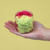 Angled view of a clear jar filled with swirls of bright pink and lime green whipped soap, held by a hand against a yellow background.