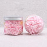 Angled view of two clear jars of pink whipped soap. The upright jar features a label with pink and yellow citrus fruit illustrations and 'Pink Lemonade Scented Whipped Soap' text, revealing the fluffy, light pink product inside. The second jar is on its side, displaying the creamy, swirled texture of the pink whipped soap.