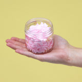 Hand holding a clear jar of pink lemonade whipped soap. The fluffy pink soap is visible, with the label showing 'Whipped Lemon Sherbet' and citrus illustrations, against a yellow background.