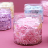 Close-up, angled view of an open clear jar filled with pink whipped soap, showing its swirled texture. The jar's label depicts citrus slices and identifies it as 'Pink Lemonade Scented Whipped Soap'.
