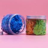 Angled view of an open clear jar of blueberry scented whipped soap, lying on its side and revealing bright blue, textured soap. Its silver-coloured lid rests detached beside it. Another lidded jar containing green and orange mango and lime whipped soap is visible in the background, all on a pink surface.