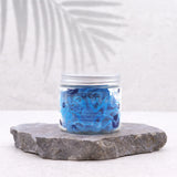 Front view of a clear jar containing light blue, swirled blueberry-scented whipped soap, with 'WHIPPED BLUEBERRY BLAST' visible on its label. The jar has a silver-coloured lid and sits on a grey stone slab, against a plain background with subtle leaf shadows.