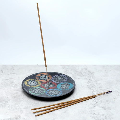 Angled view of a dark soapstone chakra incense burner dish, its top surface featuring seven colourful chakra symbols. One incense stick stands upright in the dish, with three unlit incense sticks lying nearby.