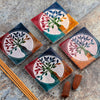 Angled overhead view of four square, multicoloured soapstone Tree of Life incense burner dishes, each featuring an engraved tree design and a central hole, displayed with brown incense sticks and cones.