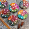 Overhead view of six round soapstone incense burner dishes, each displaying a rainbow colour gradient and intricate white carved sunburst patterns. Brown incense sticks and cones are also visible on the textured stone background.