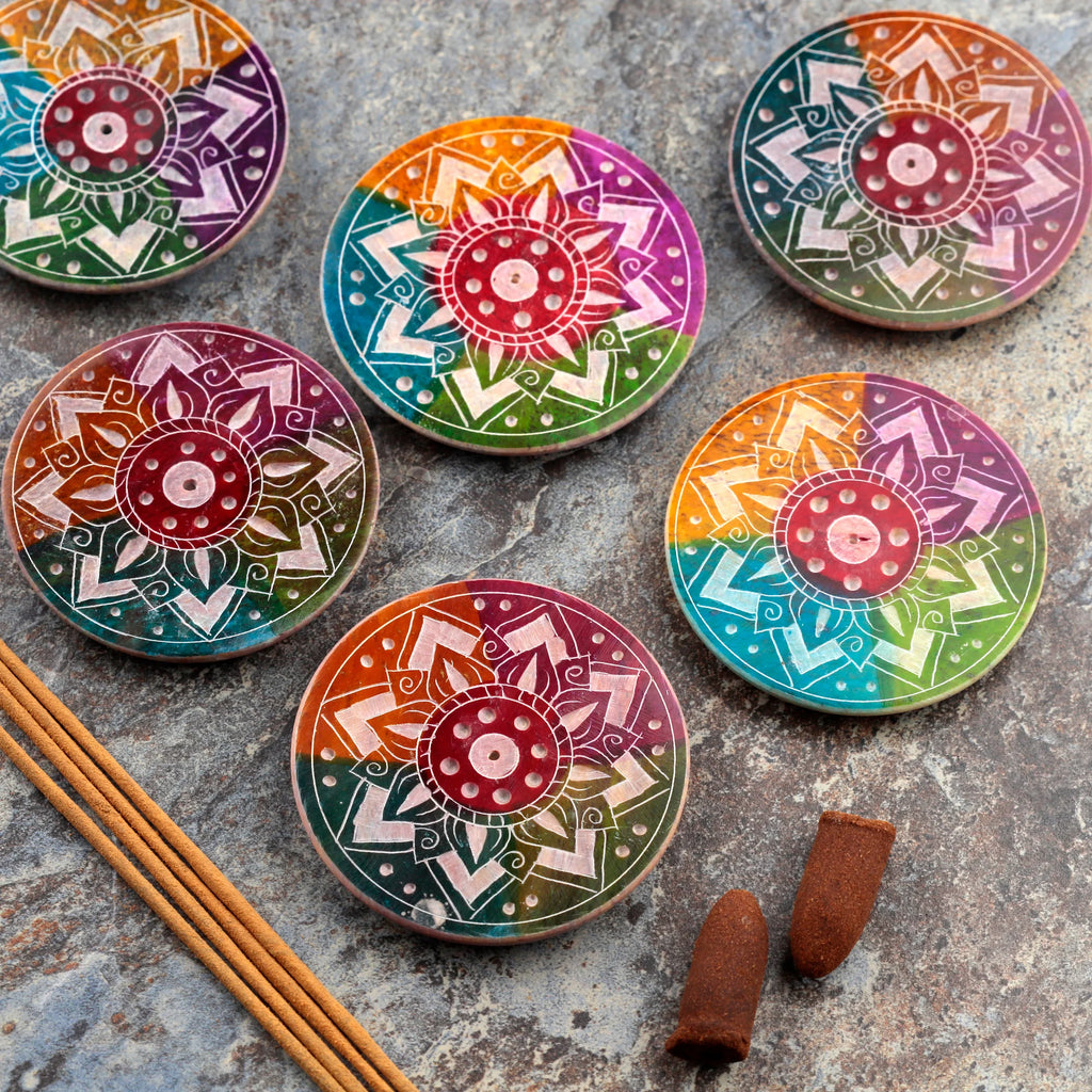 Overhead view of six round soapstone incense burner dishes, each displaying a rainbow colour gradient and intricate white carved sunburst patterns. Brown incense sticks and cones are also visible on the textured stone background.