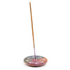 Angled view of a round soapstone incense burner dish, featuring a carved sunburst mandala pattern in reddish-brown, dark green, and purple hues, holding an upright brown incense stick.