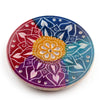 Angled top-down view of a multi-coloured soapstone incense burner dish, featuring carved white flower petal designs. It displays vibrant sections of red, purple, blue, and teal, with a yellow-orange floral centre.
