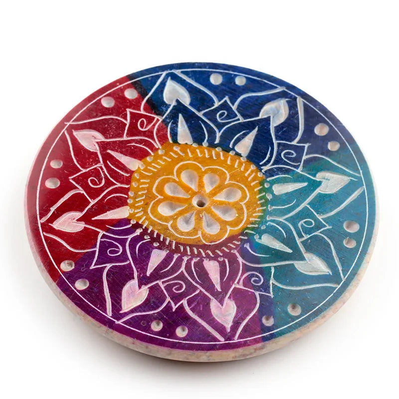 Angled top-down view of a multi-coloured soapstone incense burner dish, featuring carved white flower petal designs. It displays vibrant sections of red, purple, blue, and teal, with a yellow-orange floral centre.