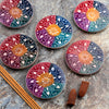 Overhead view of six vibrant multi-colour soapstone incense burner dishes, featuring white carved flower petal mandala designs, alongside brown incense sticks and cones on a grey stone surface.