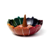 Angled view of a multi-coloured carved soapstone lotus-shaped incense burner dish, showing red, orange, green, and dark blue petals with white carved lines, and a brown incense cone in the centre.