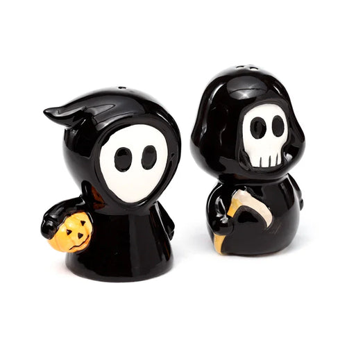 Angled view of a ceramic black and white salt and pepper set. The 'Ghost Boy' shaker, a hooded figure with an oval face, holds an orange jack-o'-lantern. The 'Skull Boy' shaker, a hooded figure with a skull face, holds a yellow scythe.