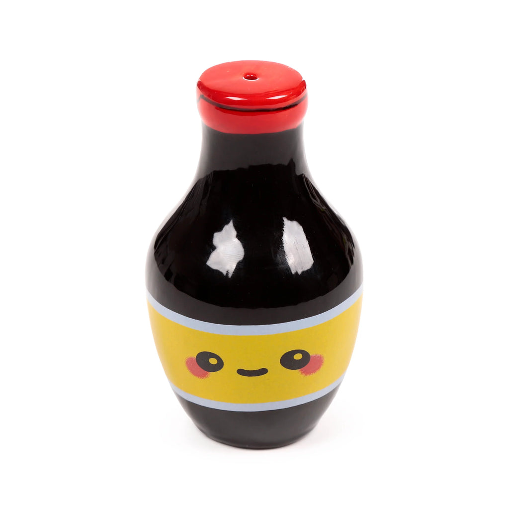 Front view of a ceramic shaker from a salt and pepper set, shaped like a glossy black soy sauce bottle. It features a yellow band with a cute cartoon face and a red cap with a single dispensing hole.